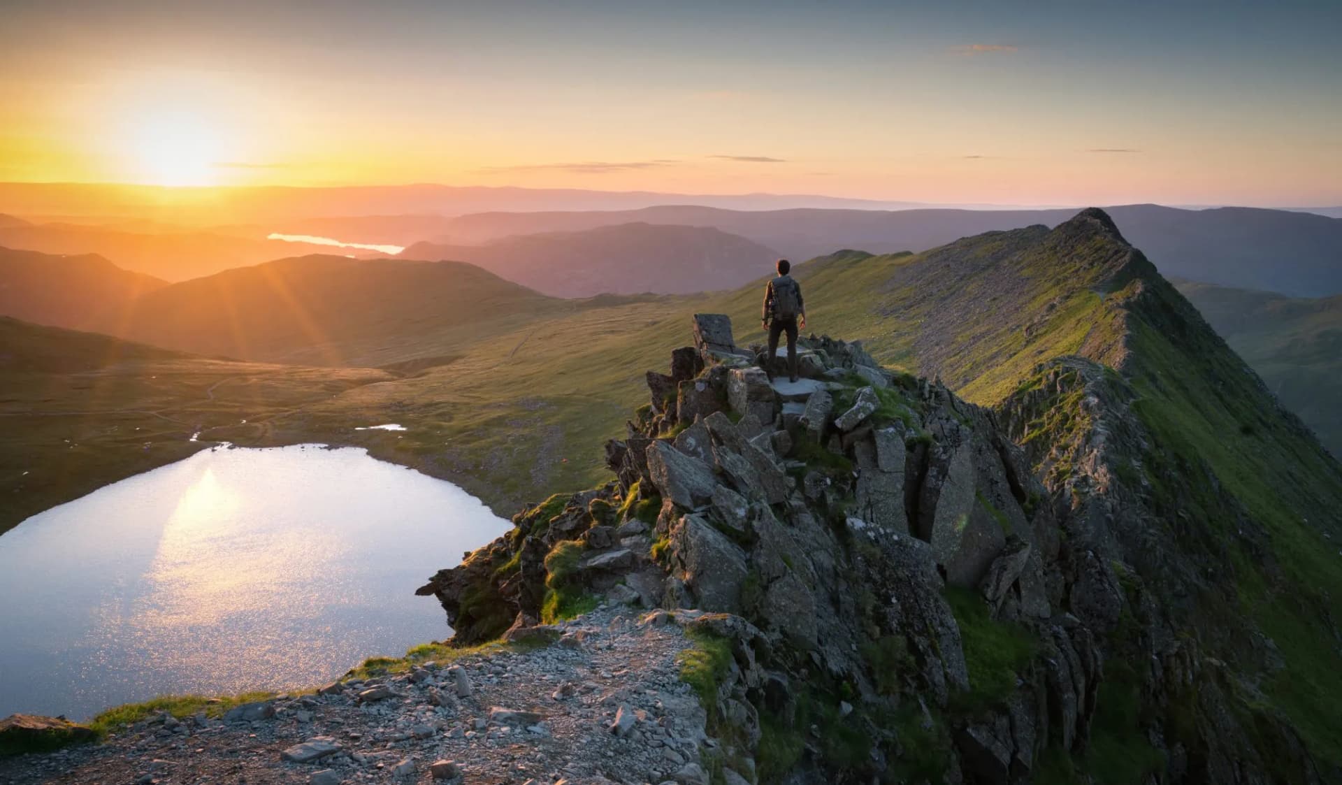 Hiker surveying the summit of Helvellyn at sunrise, with first light illuminating the rugged felltop and the tarn below